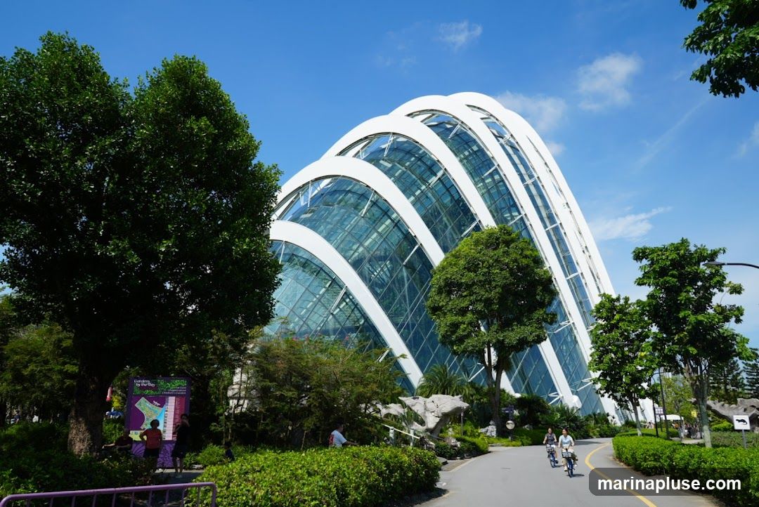 A large, arched glass conservatory with white structural elements stands under a bright blue sky with scattered clouds. Lush green trees and manicured bushes surround the building, and a paved pathway winds through the landscape. Several people are visible, some walking and others cycling, enjoying the outdoor space. A map or information board is also present near the entrance area.