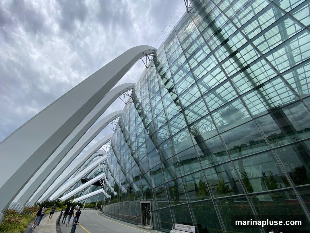 A wide-angle shot of the exterior of a large, modern conservatory with a glass facade and white arched supports. The sky is overcast. Several people are walking along a path in front of the structure.