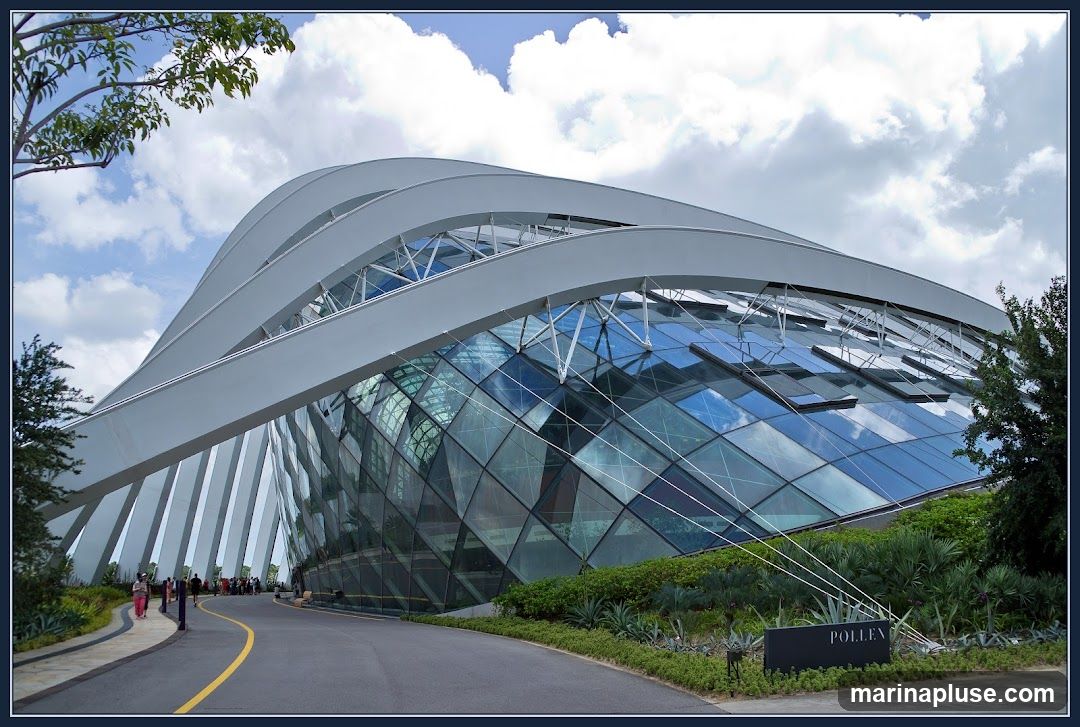 A wide shot of the exterior of the Flower Dome at Gardens by the Bay, Singapore. The dome is a large, geodesic structure with a glass facade and a white, curved roof. The building is surrounded by lush greenery and a paved walkway. Several people are visible walking along the path. A sign that reads "POLLEN" is visible in the foreground.