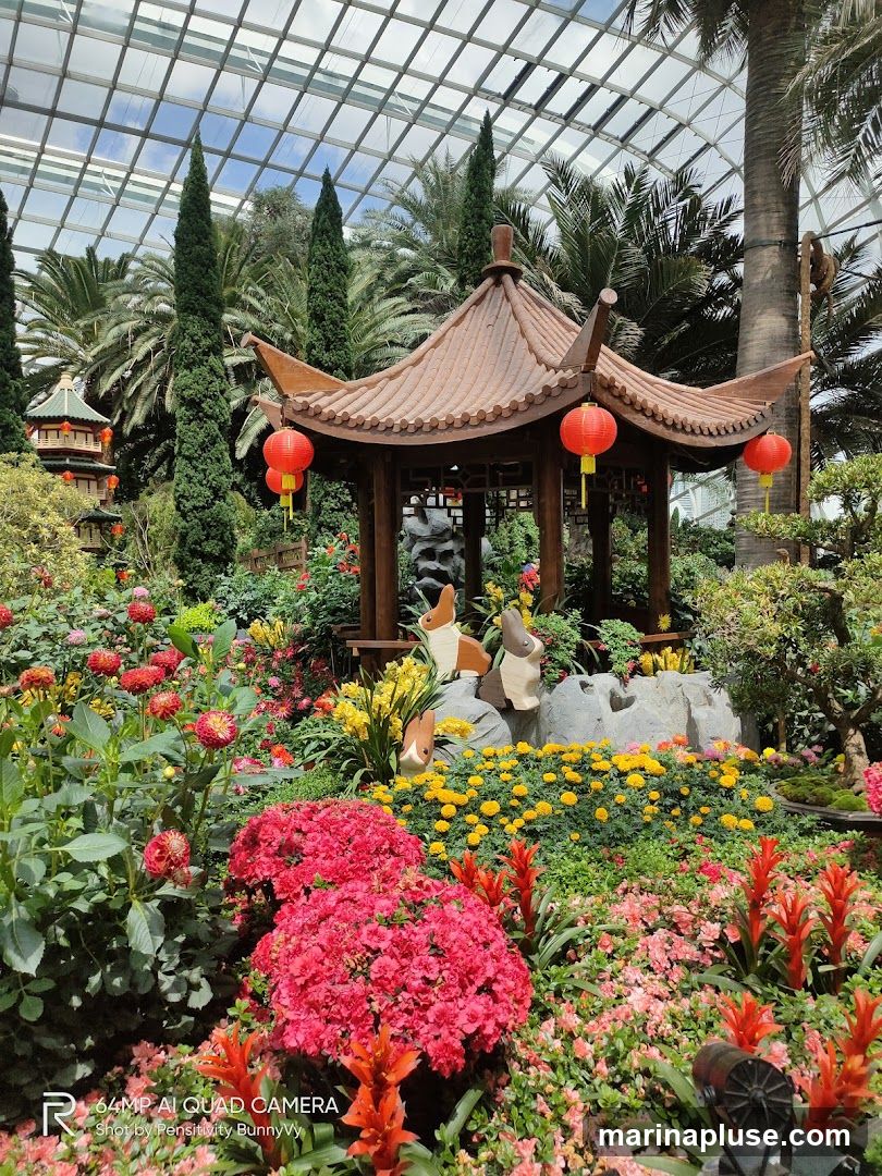 A vibrant garden display featuring a traditional Chinese pavilion, red lanterns, and an abundance of colorful flowers including red azaleas, dahlias, and bromeliads. Two wooden rabbit figurines are placed near a rock formation. The scene is set under a glass-domed ceiling, with palm trees and other foliage visible in the background.