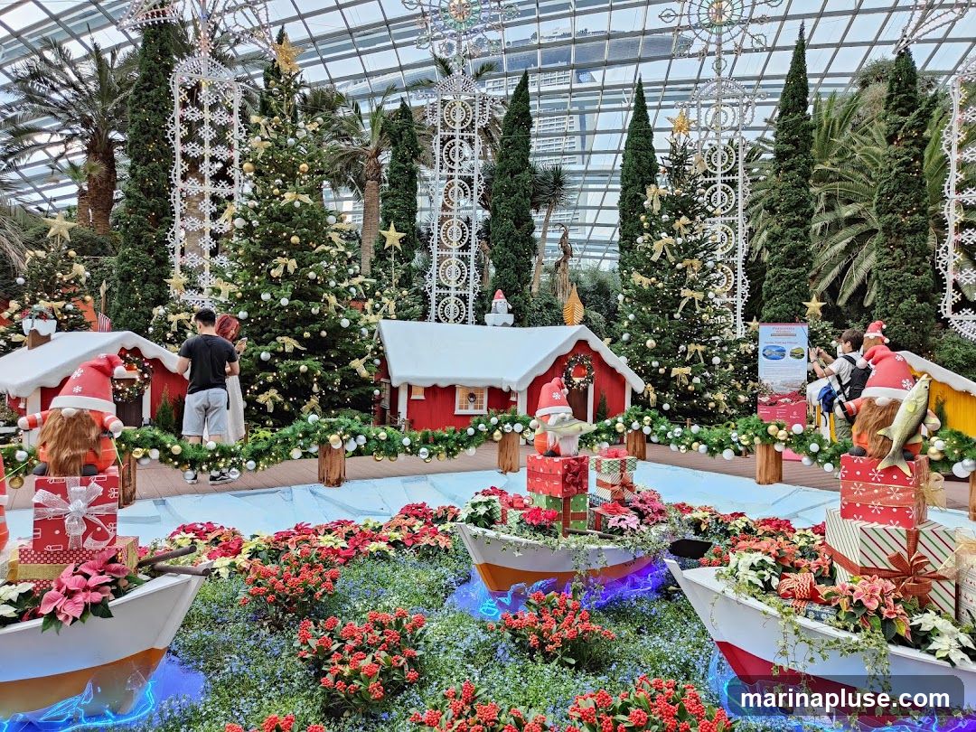 A festive Christmas display features decorated Christmas trees, miniature red houses with snow-capped roofs, and gnome figures. Boats filled with poinsettias and gift boxes are arranged in the foreground, with blue lighting effects around them. Several people are observing the display. A sign with images and text is visible in the background.