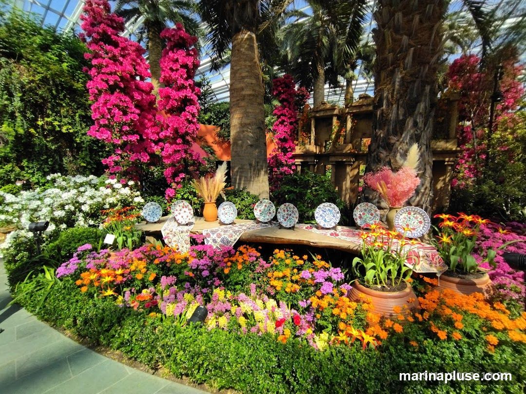 A vibrant display of flowers, including pink azaleas, white blossoms, and orange and yellow blooms, arranged around a wooden bench. Several decorative plates with intricate patterns are displayed on the bench, along with a clay vase holding dried grasses and a pink floral arrangement. In the background, palm trees and a structure resembling ancient ruins are visible, all under a glass-domed ceiling. The scene is illuminated by natural light. A date stamp in the lower right corner reads '05/11/2023 10:32'.