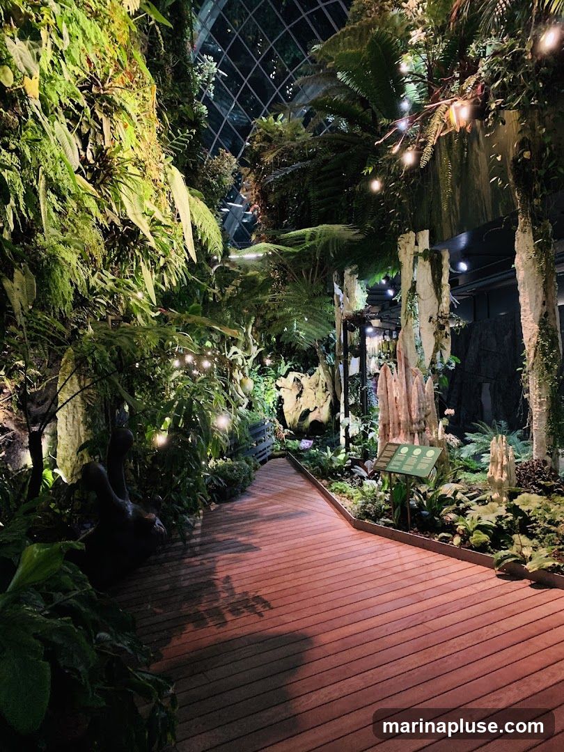 A wooden walkway winds through a lush indoor garden filled with diverse plant life, including ferns and vertical gardens. String lights are intermittently placed throughout the foliage, casting a warm glow. The scene is enclosed by a glass ceiling, suggesting a conservatory or biodome. A small informational sign is visible near the right side of the path.