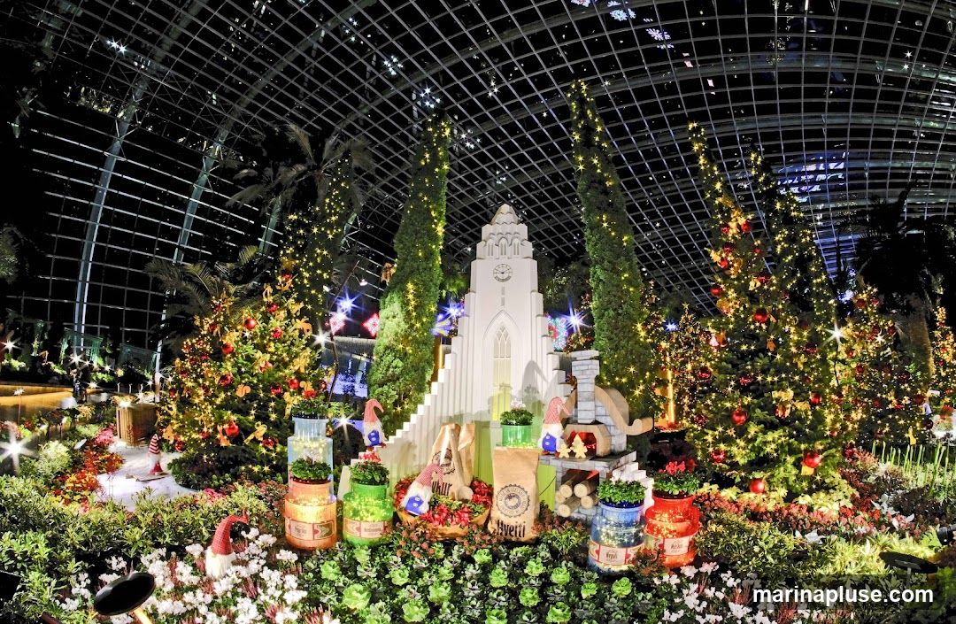 A festive Christmas display featuring decorated trees, a white church-like structure with a clock tower, and various garden gnomes. The scene is illuminated by numerous fairy lights, creating a magical atmosphere under a grid-like ceiling structure. The foreground is filled with lush greenery and flowering plants.