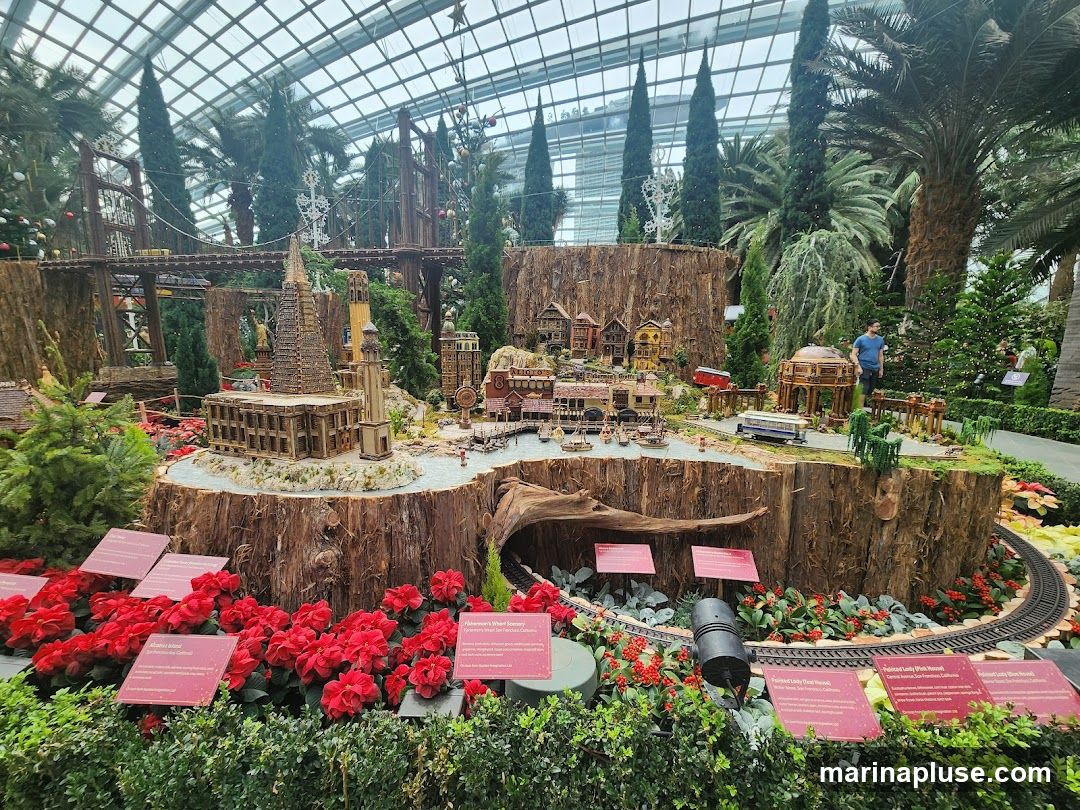 A detailed miniature village display is set up within a large greenhouse. The display features a model train track winding through a landscape with miniature buildings, bridges, trees, and a river. Red poinsettias and green foliage surround the display. Several informational signs are placed in front of the exhibit. Two people are visible in the background.