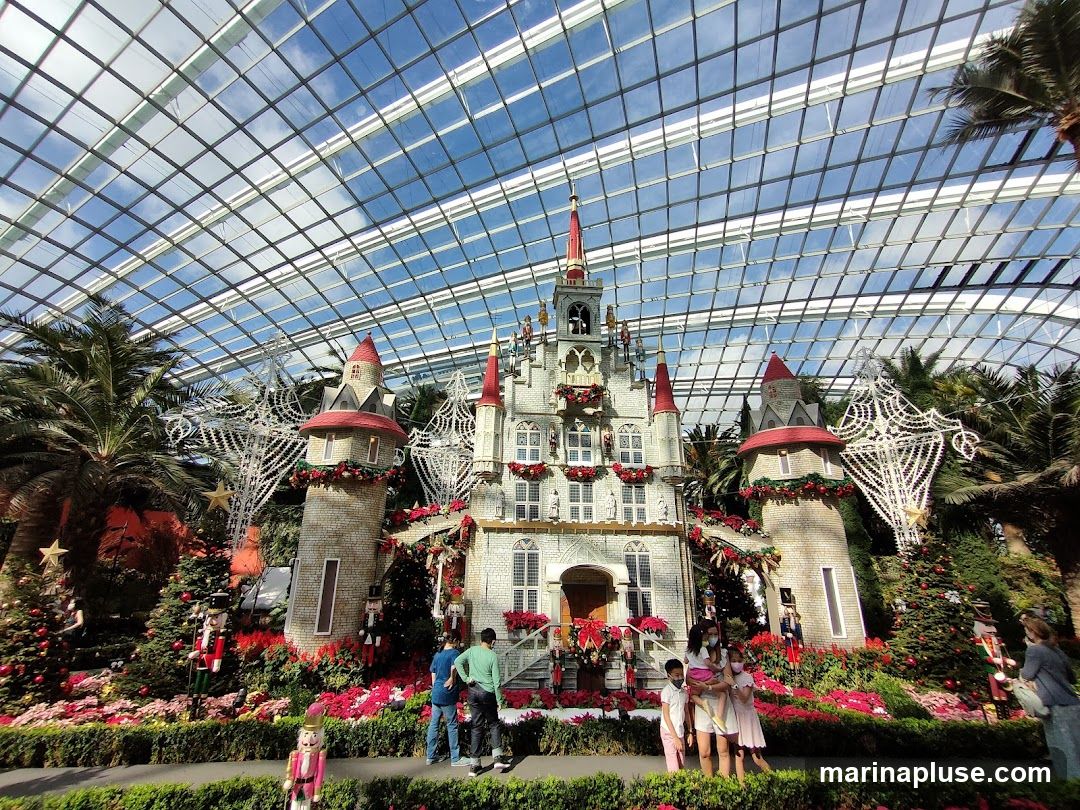 A large, ornate castle structure decorated for Christmas is displayed under a glass-paneled roof. The castle features multiple towers, red-tipped spires, and is adorned with garlands and red flowers. Surrounding the castle are Christmas trees, nutcracker figures, and lush greenery. Several people are visible, observing the display.