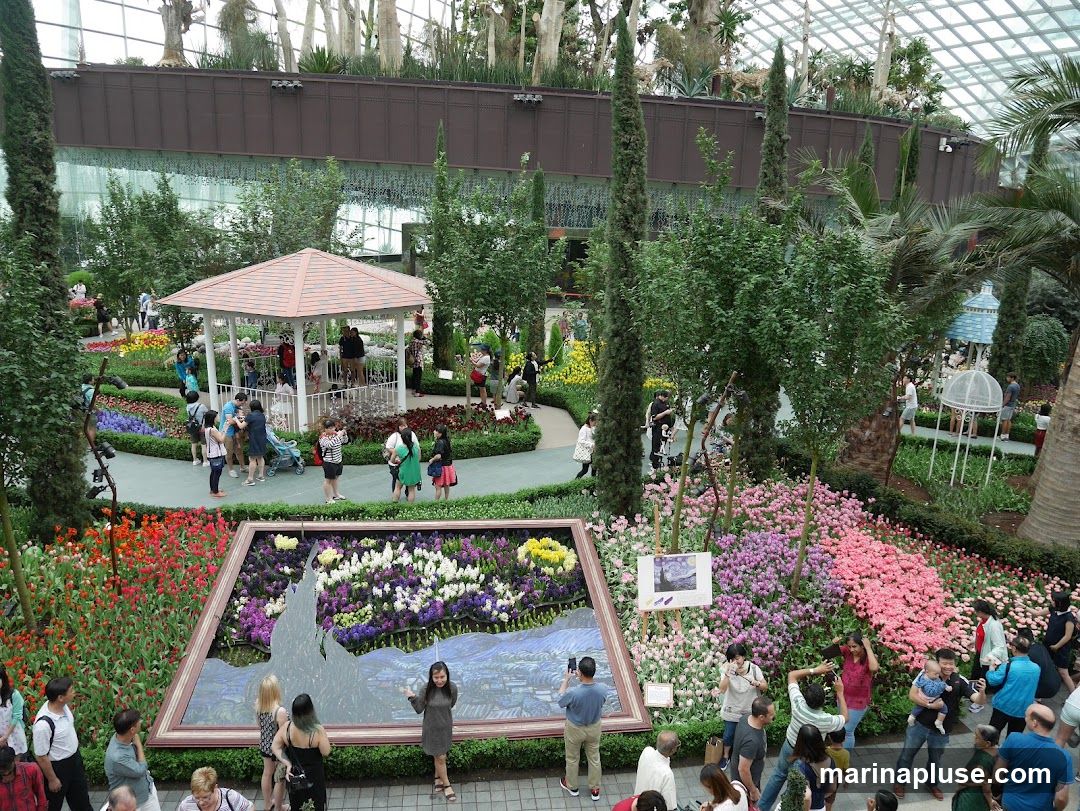 An overhead view of a vibrant flower garden with a large framed display of flowers in the foreground. People are scattered throughout the garden, admiring the floral arrangements and taking pictures. A gazebo and various trees and plants are also visible, all under a glass-domed ceiling.
