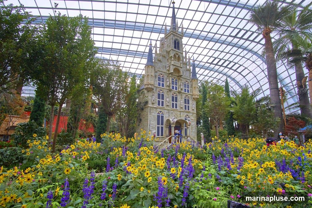 A vibrant display of yellow and purple flowers in bloom, with a miniature, ornate building resembling a European-style structure in the background. The scene is enclosed by a glass ceiling, suggesting an indoor botanical garden or conservatory. Palm trees and other greenery are also visible, contributing to the lush atmosphere.