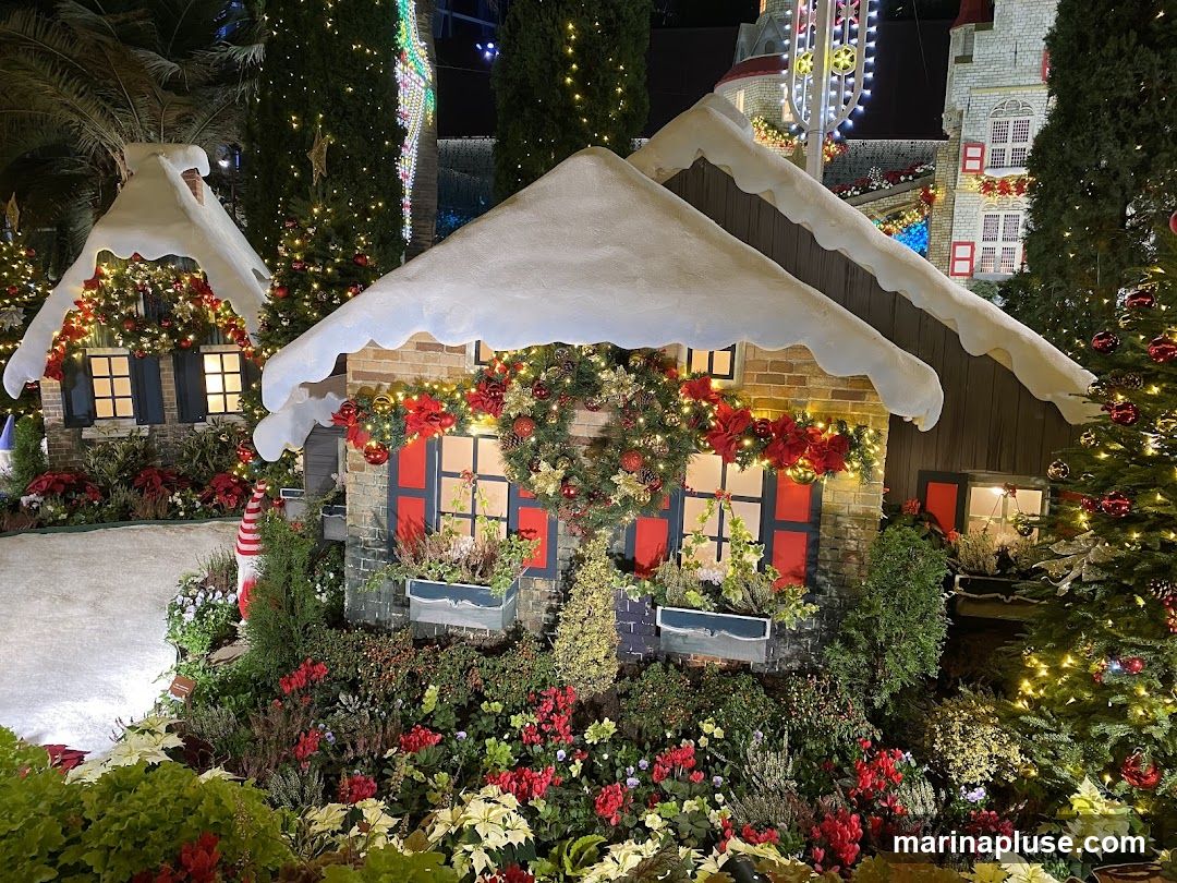 A festive display of miniature houses decorated for Christmas, surrounded by lush greenery and poinsettias. The houses have snow-covered roofs and are adorned with wreaths, garlands, and twinkling lights. A Christmas tree with red ornaments stands to the right. In the background, larger buildings and a Ferris wheel-like structure are visible, also decorated with lights.
