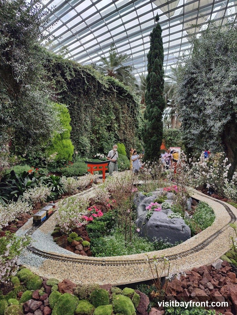 A miniature train track winds through a lush garden display under a glass-domed ceiling. The garden features a variety of plants, including flowering trees, bushes, and moss-covered rocks. A small red torii gate is visible in the background, and several people are observing the display.