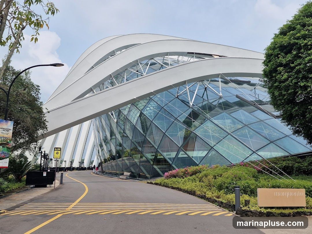 A wide shot of the exterior of a large, modern conservatory building with a distinctive curved white roof and a faceted glass facade. A paved pathway leads towards the entrance, bordered by lush greenery and flowering plants. A sign with the word "marquente" is visible on the right. A poster advertising "Impressions of Monet" is on the left.