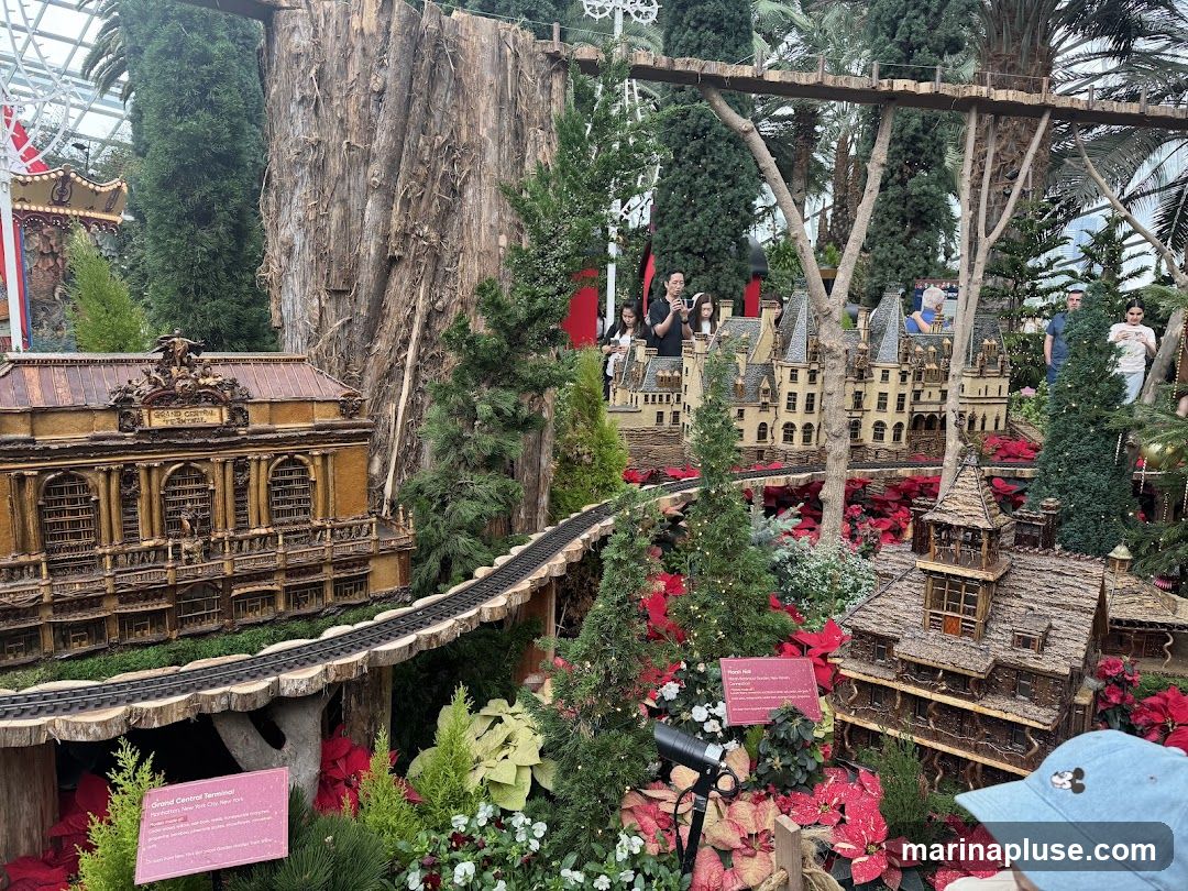 A miniature train display featuring detailed models of buildings and landscapes, set amongst lush greenery and poinsettias. A train track winds through the scene, connecting various structures. People are visible in the background observing the display.
