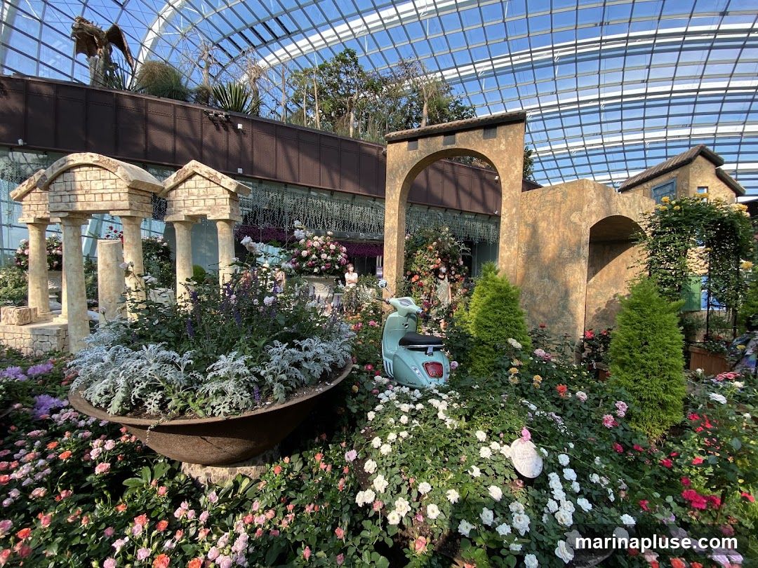 A vibrant floral display featuring numerous roses in various shades of pink, white, and orange, interspersed with greenery. In the background, Roman-inspired architectural elements, including columns and arches, are visible, along with a light blue vintage scooter. The scene is set within a large greenhouse with a glass ceiling.