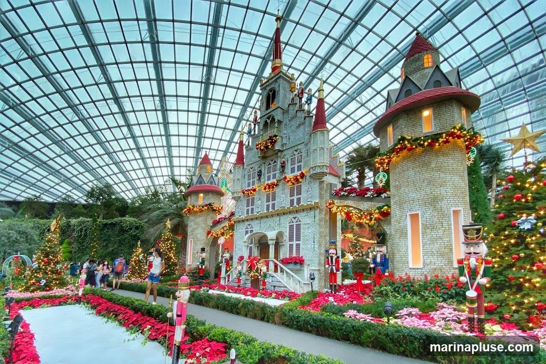 A festive Christmas display featuring a large, elaborate castle structure adorned with lights and garlands. Nutcracker figures stand guard along a pathway lined with poinsettias and Christmas trees. People are visible in the background, enjoying the holiday decorations within a large, glass-enclosed space.