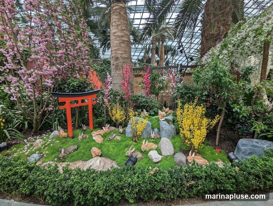 A vibrant display of a Japanese-inspired garden featuring a bright orange torii gate, cherry blossom trees with pink flowers, yellow flowering bushes, and various green foliage. Numerous rabbit figurines are scattered throughout the mossy ground, some resting on rocks. Palm trees and a glass ceiling are visible in the background, suggesting an indoor botanical garden setting.