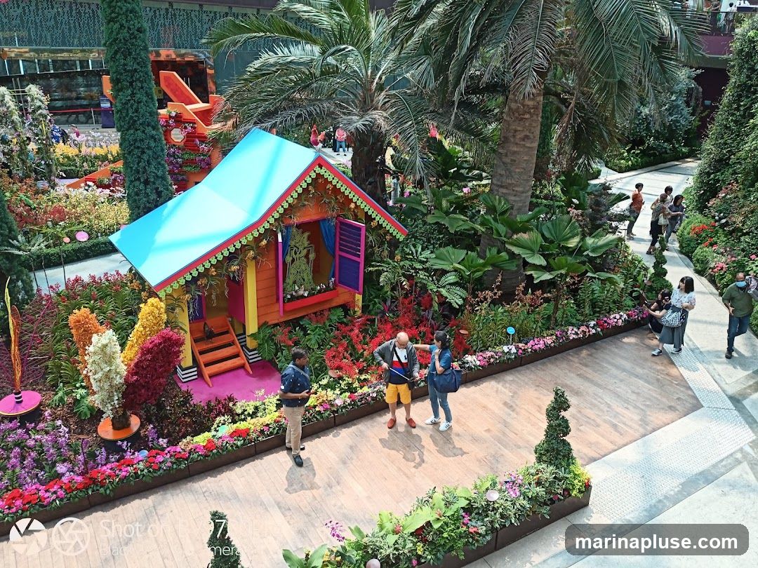 A vibrant and colorful miniature house is the centerpiece of a lush floral display. The house, painted in bright yellow, pink, and blue, features an open doorway with stairs leading up to it. Inside, a traditional puppet show scene is set up. The house is surrounded by an abundance of flowers in various colors, including red, orange, yellow, white, and purple, as well as lush green foliage and palm trees. Several people are seen walking around the display, admiring the scenery.