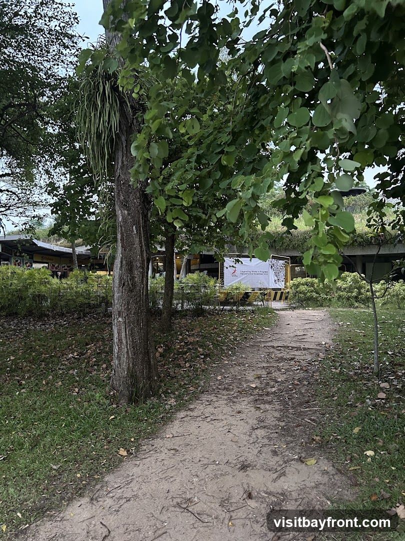 A dirt path winds through a grassy area with trees and fallen leaves. In the background, there is a building with a green roof and a yellow sign with text and graphics.