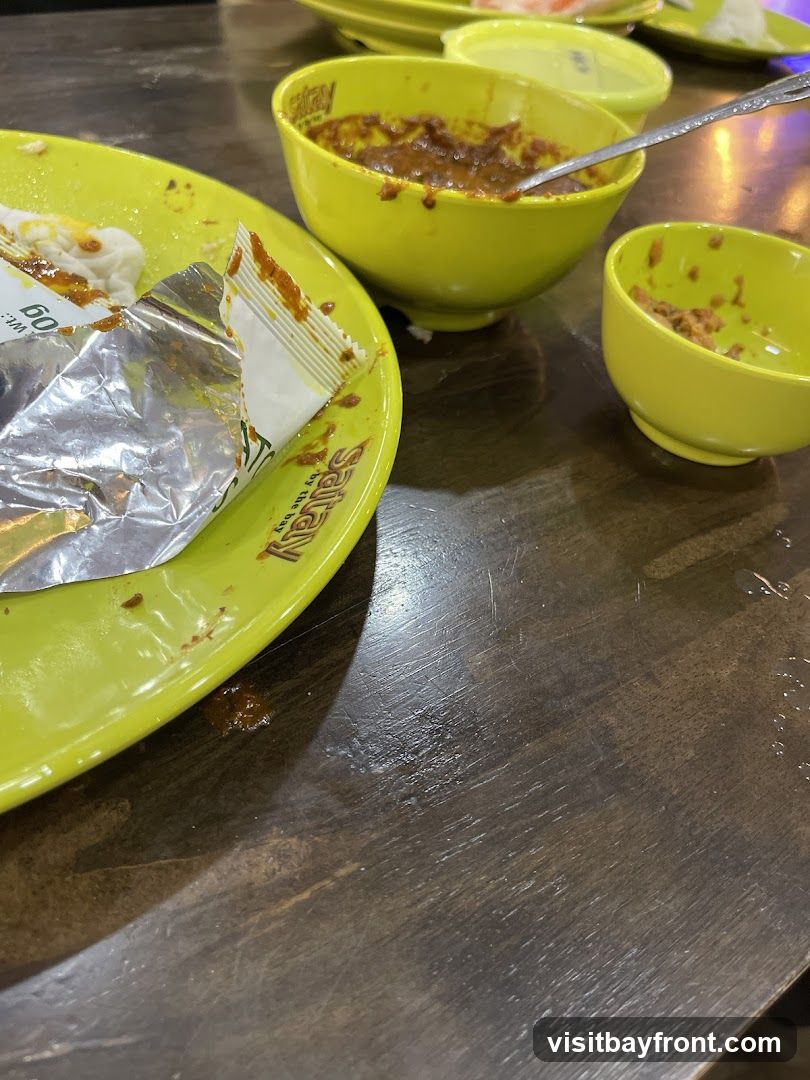 A close-up view of a table with leftover food and dishes. A bright green plate with a foil wrapper and some food residue is in the foreground. To the right, two bright green bowls contain remnants of a dark, saucy dish. A spoon is visible in the larger bowl. The table surface is dark wood with visible scratches.