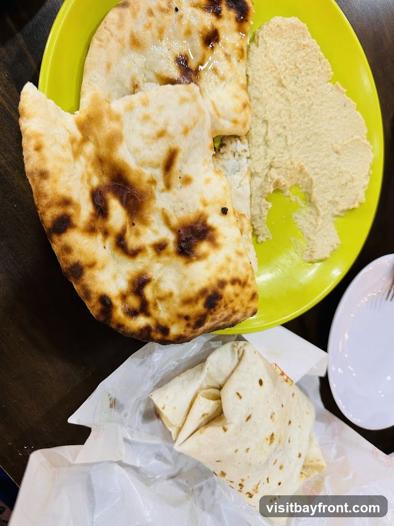 A close-up overhead shot shows a bright yellow plate filled with pieces of warm naan bread and a serving of hummus. Below the plate, a wrap is presented in white paper packaging. A white plate with a fork is visible to the right.