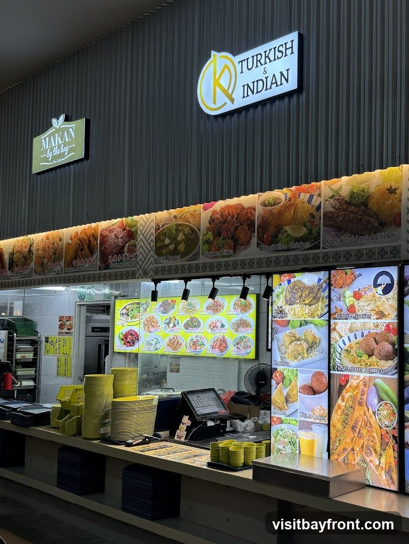 A food stall with signs for "MAKAN by the bag" and "TURKISH & INDIAN". Displayed are various food items including Mandi Rice Chicken, Adana Kebab Lamb & Chicken, Butter Chicken, and Falafel. The stall has a counter with stacks of plates and bowls, a cash register, and a fan.