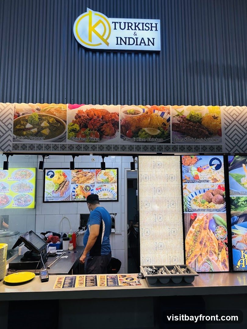 A food stall with a sign reading "TURKISH & INDIAN" above a display of food images. The images show dishes like Palak Paneer, Chicken Tikka, Mandi Rice Chicken, Adana Kebab Lamb & Chicken, Butter Chicken, and Falafel. A staff member is visible behind the counter.
