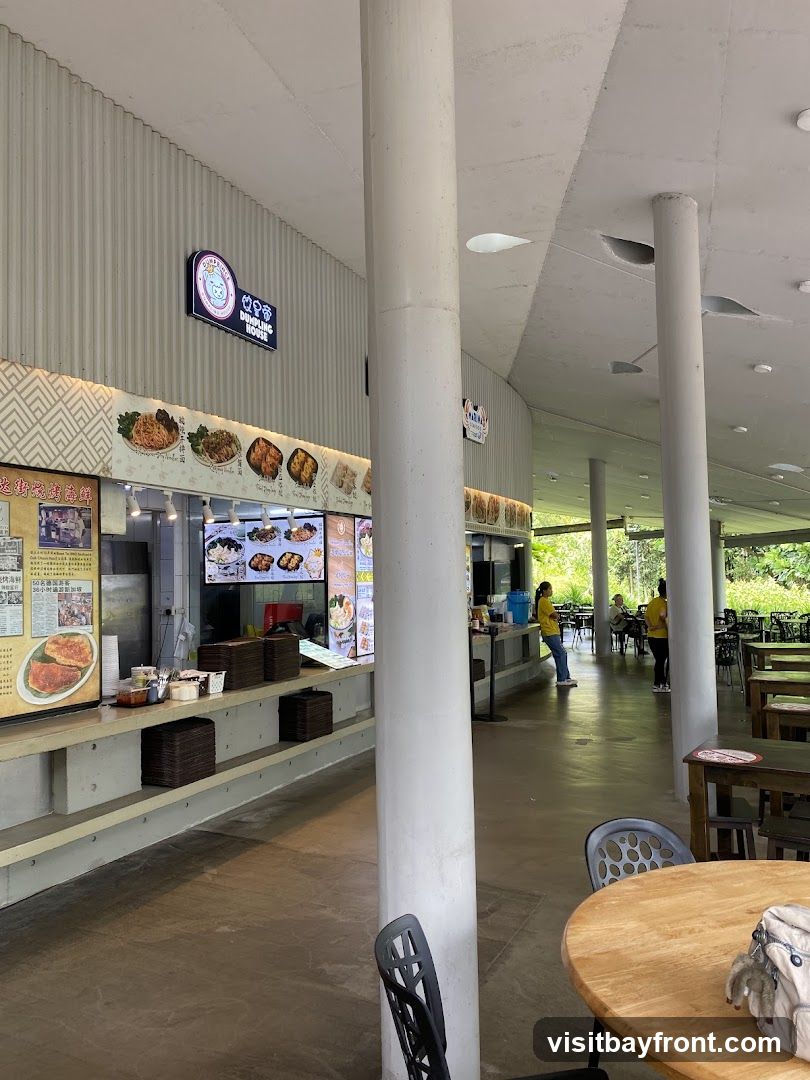 An open-air food court with multiple food stalls and seating areas. One stall,