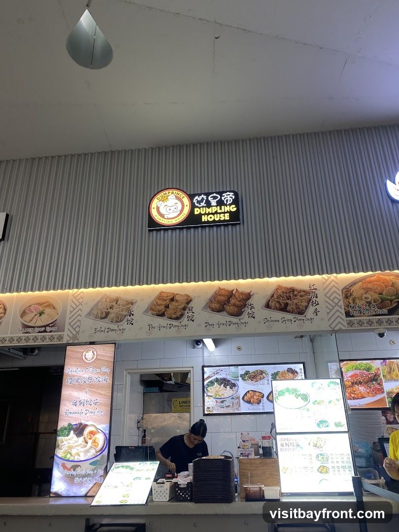 A food stall counter with a sign that reads "DUMPRINCE DUMPLING HOUSE". There are various food pictures displayed on the wall and digital screens. A staff member is seen preparing food behind the counter.