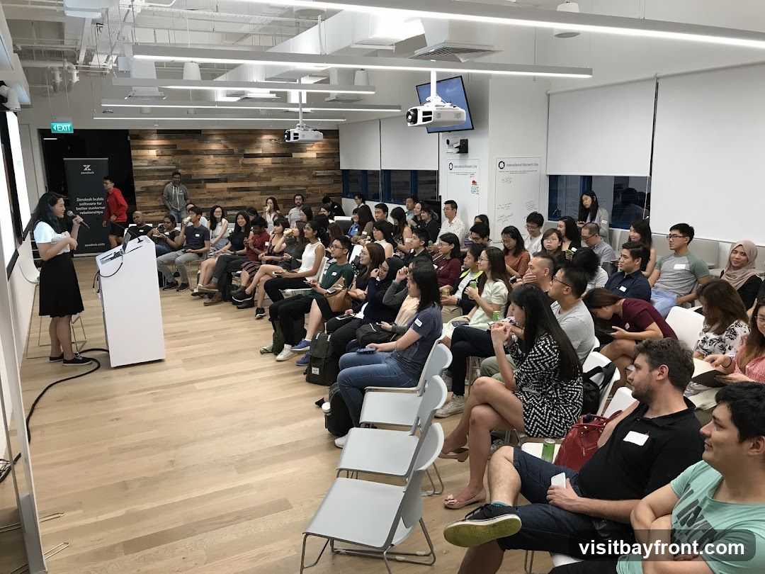 A woman is presenting to a seated audience in a bright, modern office space. The audience is diverse and appears engaged. The room has a wooden accent wall, whiteboards with writing, and a projector mounted on the ceiling. There is a large screen on the left side of the room, and a podium with a microphone in front of the presenter.