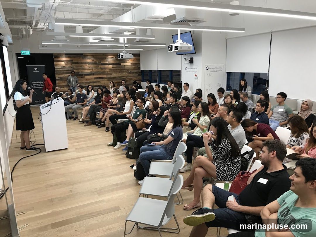 A woman is presenting to a seated audience in a bright, modern office space. The audience is diverse and appears engaged. The room has a wooden accent wall, whiteboards with writing, and a projector mounted on the ceiling. There is a large screen on the left side of the room, and a podium with a microphone in front of the presenter.