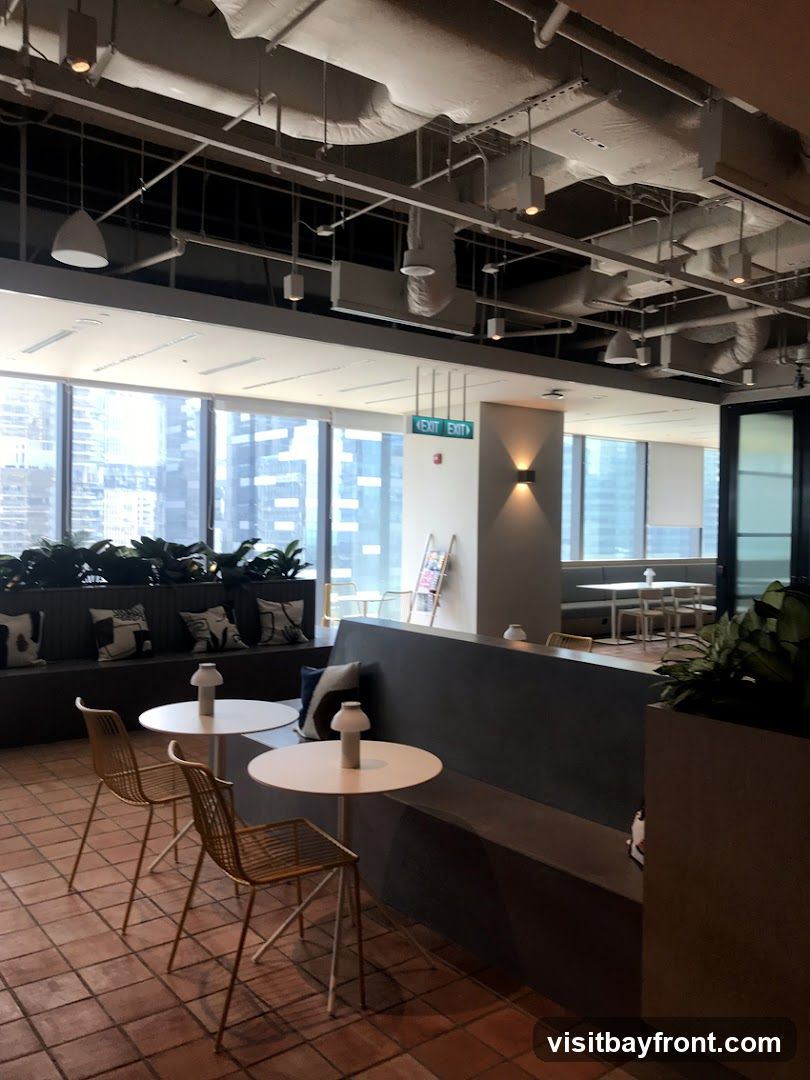 An interior view of a modern office break area with large windows overlooking a city skyline. The space features a long, dark gray bench seating area with decorative pillows, two small white circular tables with minimalist lamps, and gold wire chairs. Exposed ceiling pipes and industrial-style lighting add to the contemporary aesthetic. Green plants are strategically placed throughout the area. An illuminated green 'EXIT' sign is visible on the wall.