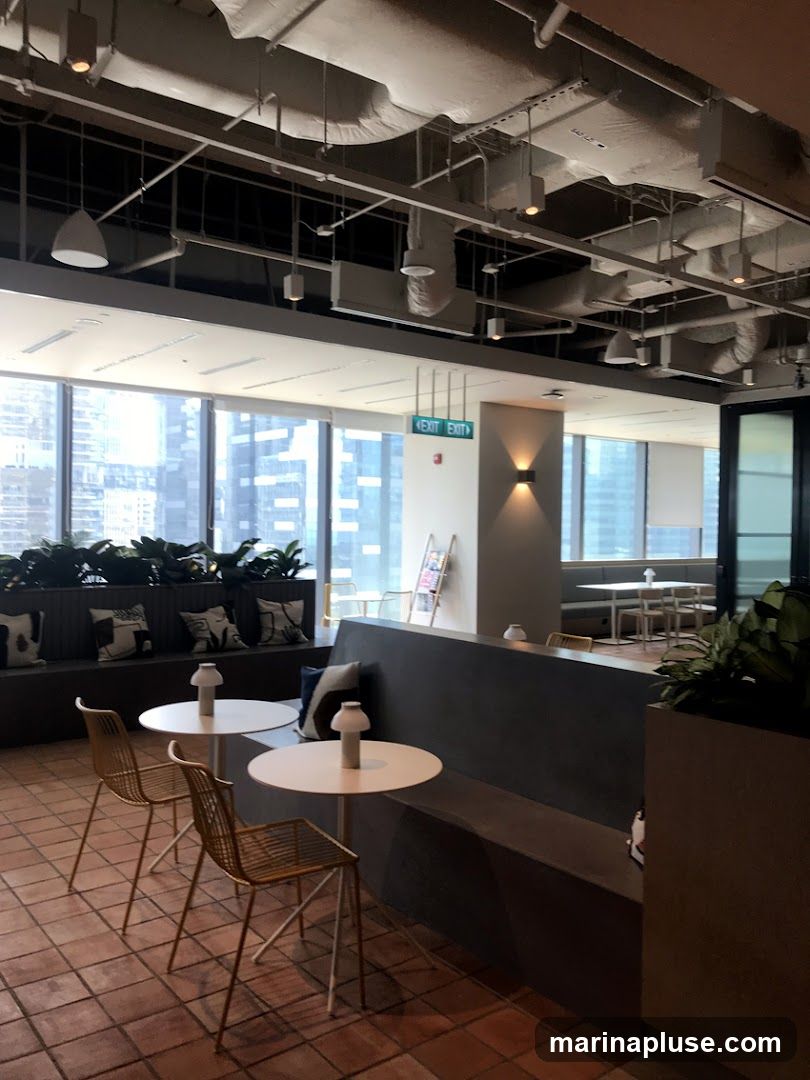 An interior view of a modern office break area with large windows overlooking a city skyline. The space features a long, dark gray bench seating area with decorative pillows, two small white circular tables with minimalist lamps, and gold wire chairs. Exposed ceiling pipes and industrial-style lighting add to the contemporary aesthetic. Green plants are strategically placed throughout the area. An illuminated green 'EXIT' sign is visible on the wall.