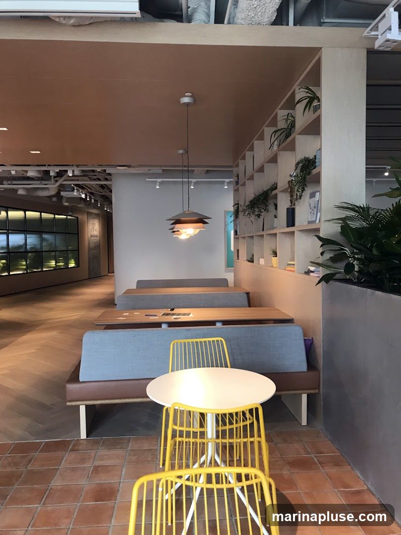 A contemporary cafe interior with booth seating and yellow chairs. The space features a wooden ceiling, a tiered pendant light, and a wall with shelves displaying plants and books. The flooring is a mix of herringbone wood and terracotta tiles.