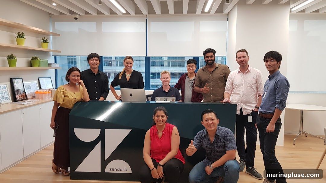 A group of nine people, appearing to be employees, are posing for a photo in an office setting. They are standing and crouching behind a reception desk with the Zendesk logo. The office appears modern with light-colored walls, wooden floors, and large windows. Shelves with plants and framed items are visible on the left side of the image.