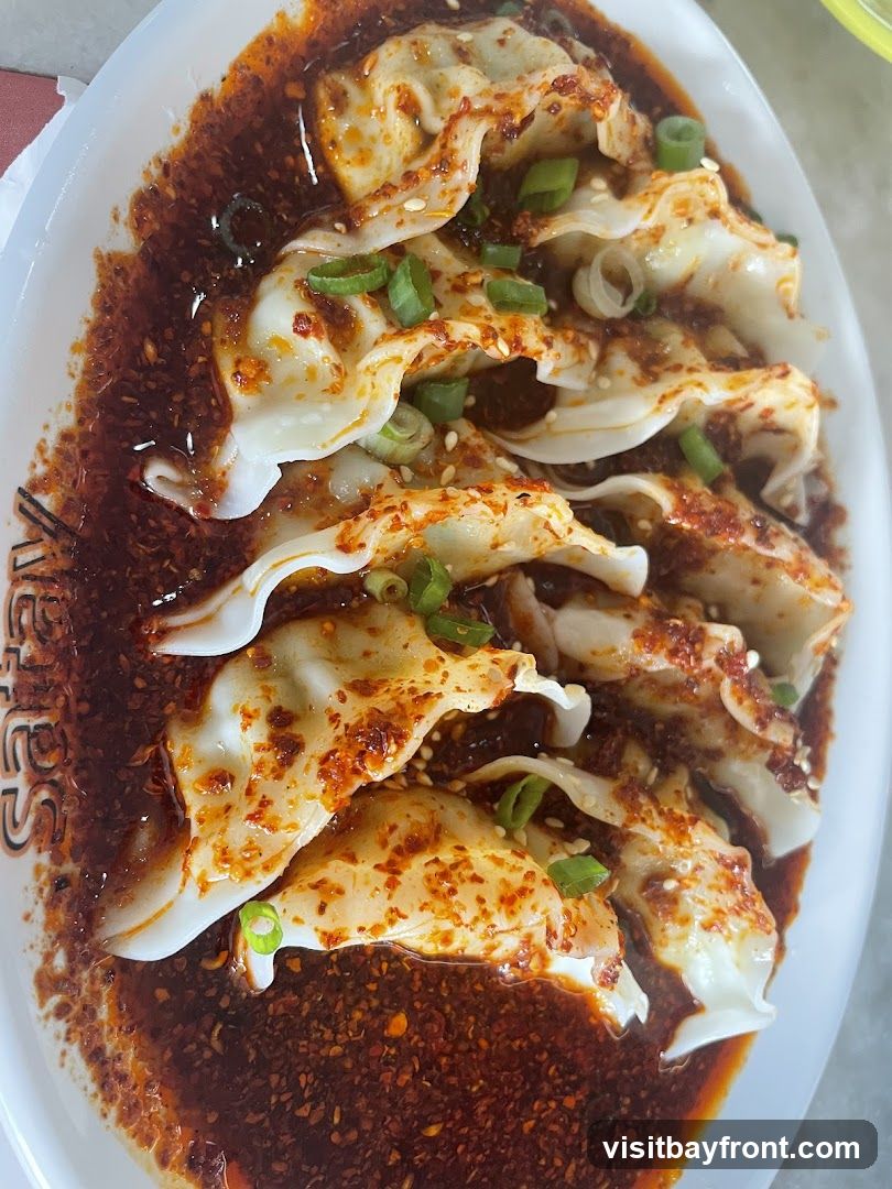 A close-up shot of several dumplings in a dark, spicy sauce, garnished with chopped green onions and sesame seeds. The dumplings are white and appear to be filled. The sauce is rich and contains chili flakes. Part of a white plate with text is visible on the left side.