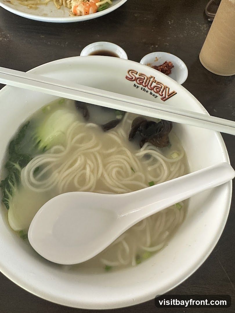 A bowl of noodle soup with bok choy and mushrooms, served with chopsticks and a spoon. The bowl has the logo "satay by the bay" printed on it. There are small dishes of condiments and a drink in the background.