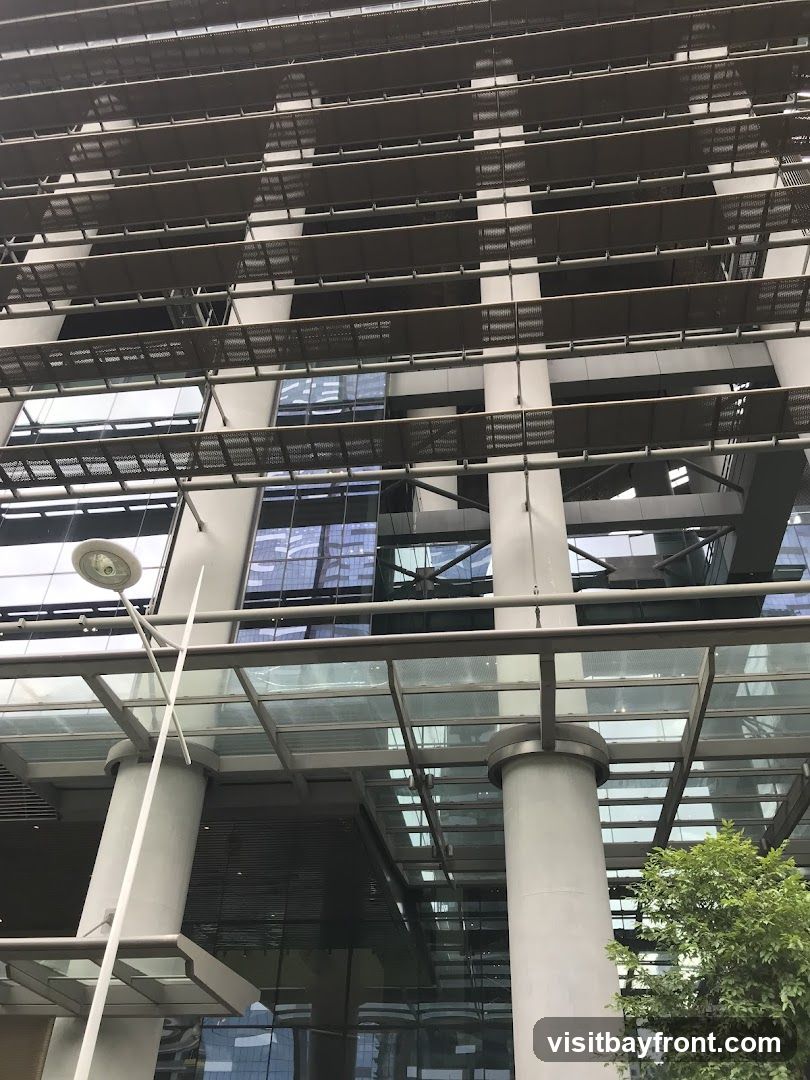 A low-angle shot of a modern building with a glass and metal facade. The building features horizontal louvers or sunshades on multiple levels, creating a layered effect. Large glass windows reflect the sky and surrounding structures. Several large, cylindrical columns support an overhead glass canopy. A street lamp is visible on the left side of the frame, and some greenery from a tree is seen on the right.