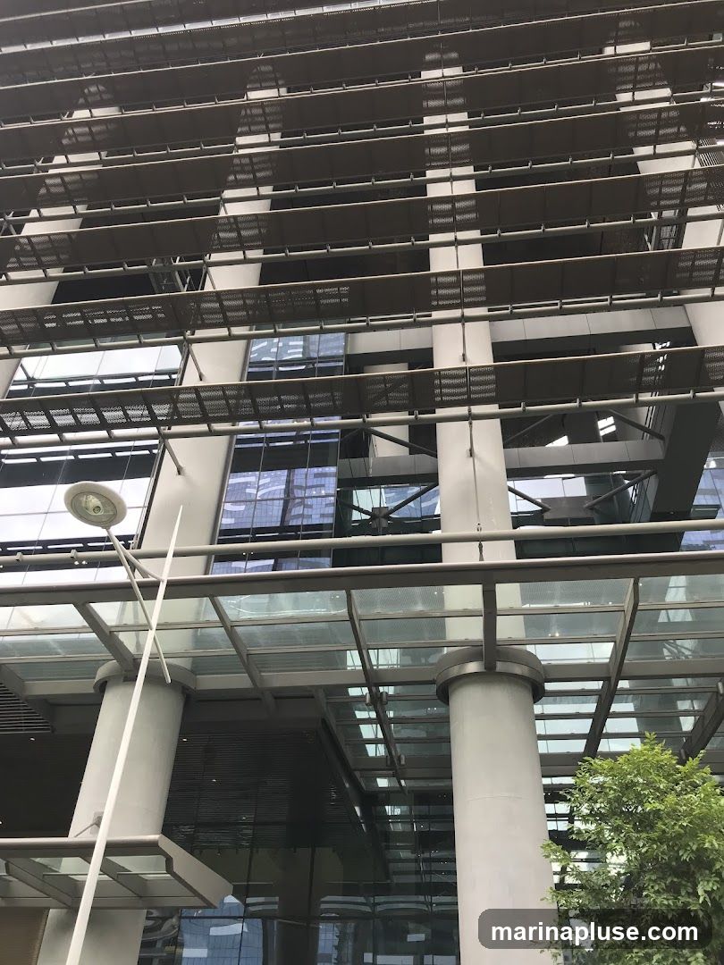 A low-angle shot of a modern building with a glass and metal facade. The building features horizontal louvers or sunshades on multiple levels, creating a layered effect. Large glass windows reflect the sky and surrounding structures. Several large, cylindrical columns support an overhead glass canopy. A street lamp is visible on the left side of the frame, and some greenery from a tree is seen on the right.