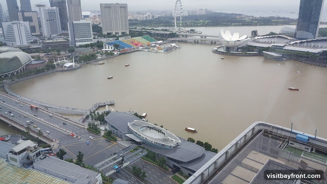 An aerial view of Marina Bay in Singapore, showcasing the city skyline, the bay with several boats, and various architectural landmarks. The image captures the Fullerton Bay Hotel, the ArtScience Museum, and the Singapore Flyer in the distance. The bay is a murky brown color, and the sky is overcast.