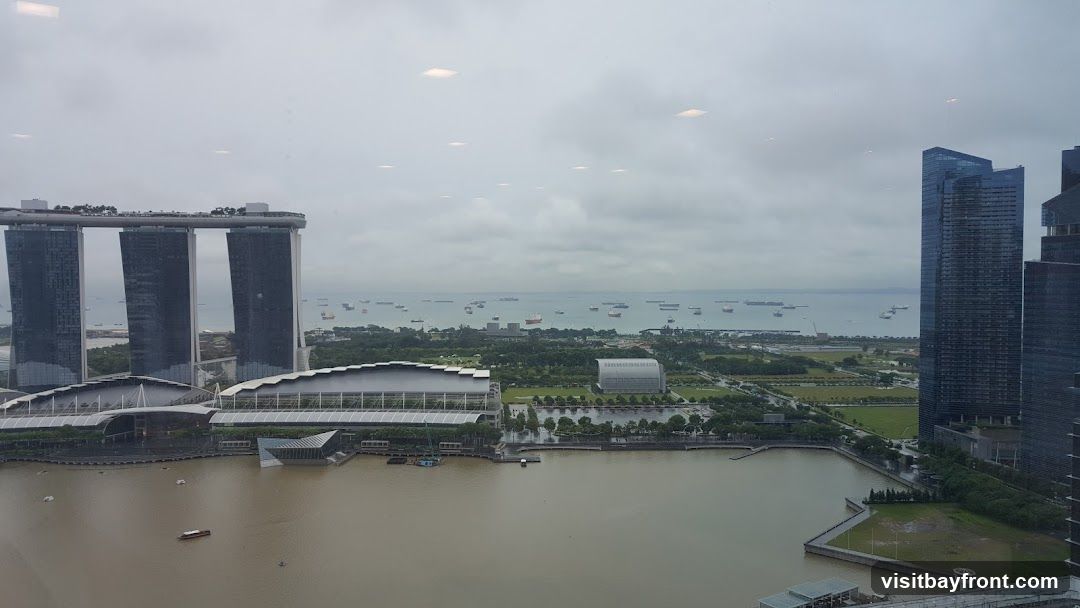 An aerial view of the Singapore skyline on a cloudy day, featuring the iconic Marina Bay Sands hotel, a large body of water, and numerous ships in the distance. Modern skyscrapers are visible on the right side of the frame.