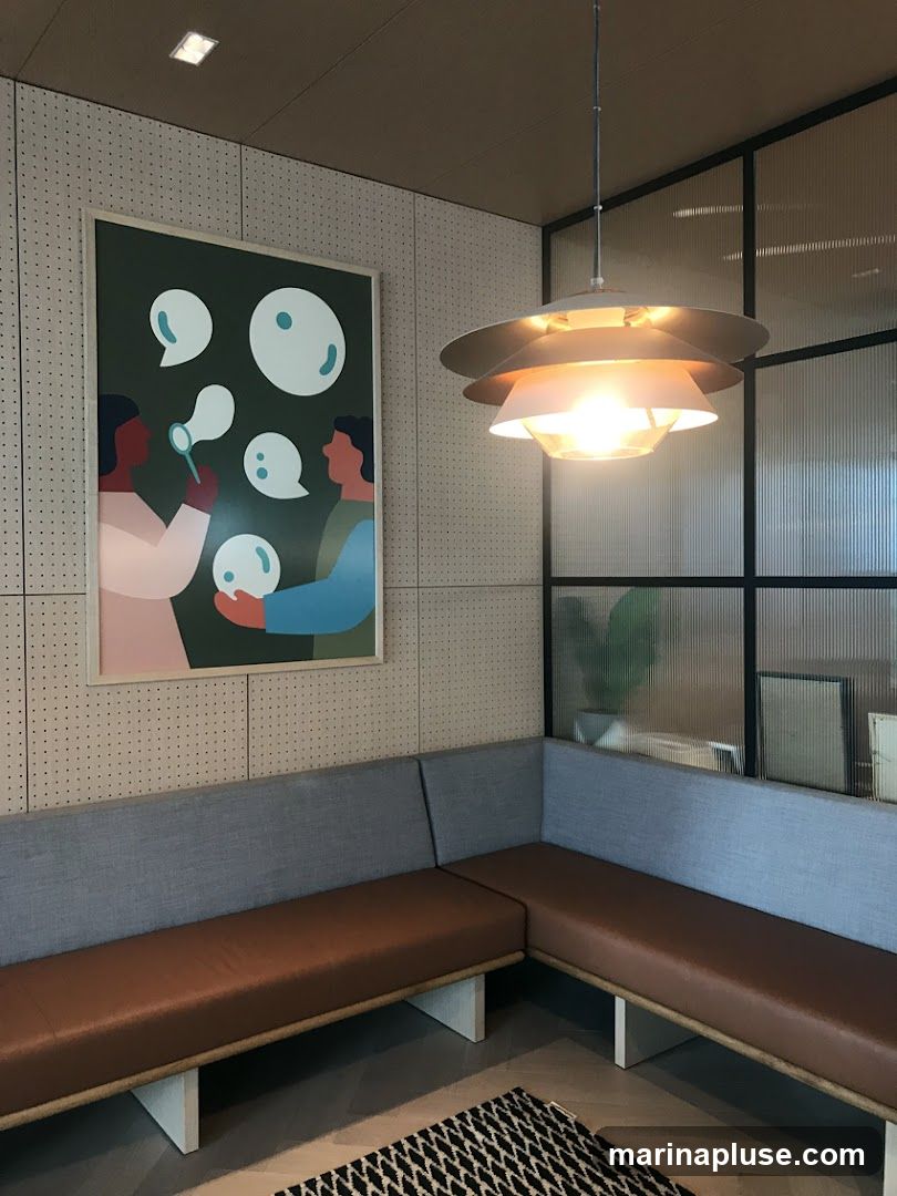 A modern waiting area features a large framed artwork depicting two figures blowing bubbles. The artwork is hung on a pegboard-style wall. A contemporary pendant light hangs from the ceiling above a sectional sofa with brown leather seats and grey fabric backs. The sofa is arranged in an L-shape. A patterned rug is visible on the floor. In the background, a glass partition with vertical lines separates the area from another space where a potted plant and framed pictures can be seen.