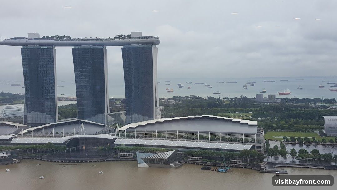 An aerial view of the Marina Bay Sands hotel in Singapore, with its iconic three towers and rooftop infinity pool. The hotel is situated on the waterfront, with a large convention center and shopping mall in the foreground. The bay is filled with numerous cargo ships, indicating a busy port. The sky is overcast, casting a muted light over the scene.