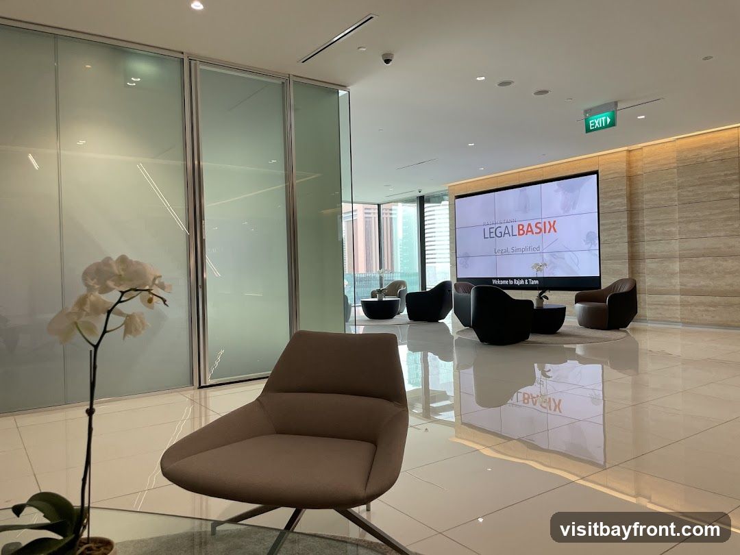 A modern and spacious office lobby with a large screen displaying the "LEGALBASIX" logo and tagline "Legal, Simplified." The screen also shows a welcome message to "Rajah & Tann." The lobby features comfortable seating areas with armchairs and coffee tables, a decorative orchid plant, and polished tiled floors that reflect the surroundings. An exit sign is visible on the ceiling.