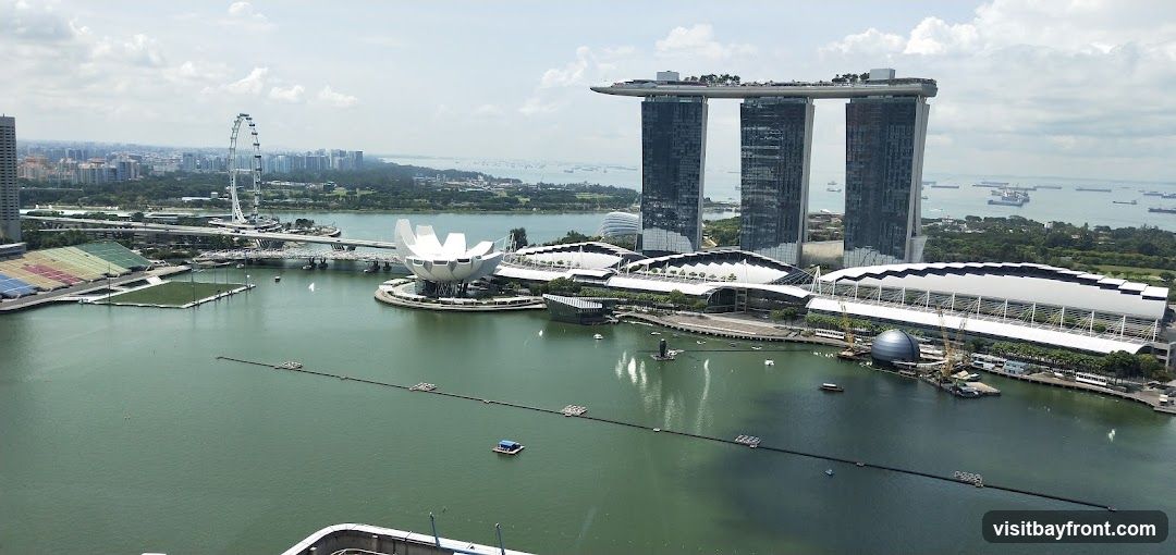 An aerial view of the Marina Bay Sands complex in Singapore, featuring its iconic three towers topped with a sky park. The ArtScience Museum, shaped like a lotus flower, is visible in the foreground. The Singapore Flyer Ferris wheel can be seen in the distance, along with a sprawling cityscape and numerous ships in the bay. The water in the bay is a murky green, and the sky is partly cloudy.