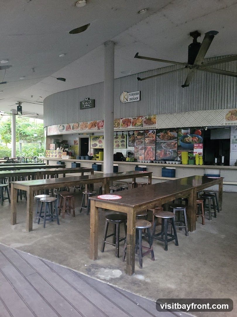 An outdoor food court with long communal tables and stools. Various food stalls are visible in the background with pictures of their offerings. Signs indicate "Makan" and "Turkish Indian" cuisine.