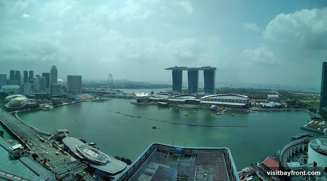 A wide-angle, panoramic view of the Singapore skyline on a hazy day. The image captures the Marina Bay area with its iconic landmarks, including the Marina Bay Sands hotel, the ArtScience Museum, and the Singapore Flyer Ferris wheel. The cityscape is dominated by modern skyscrapers and a large body of water, likely Marina Bay itself. Several boats are visible on the water, and a bridge with traffic can be seen in the foreground. The overall impression is one of a bustling, contemporary urban environment.