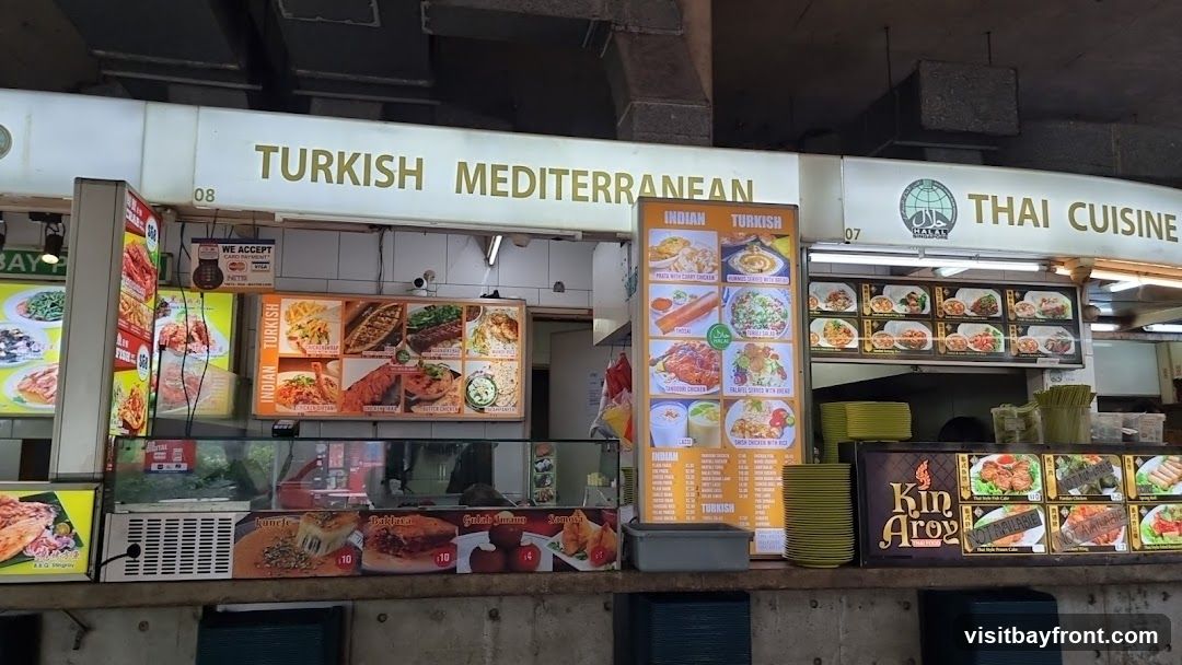 A row of food stalls with signs for Turkish, Mediterranean, Indian, and Thai cuisine. Various food items are displayed on menus and in display cases.