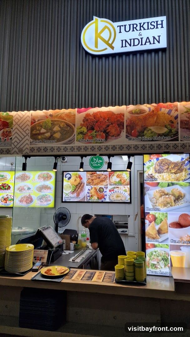 A food stall with a sign that reads "K TURKISH & INDIAN". The stall displays various food images, including Palak Paneer, Chicken Tikka, Mandi Rice Chicken, Biryani, and Baklava. A staff member is visible behind the counter.