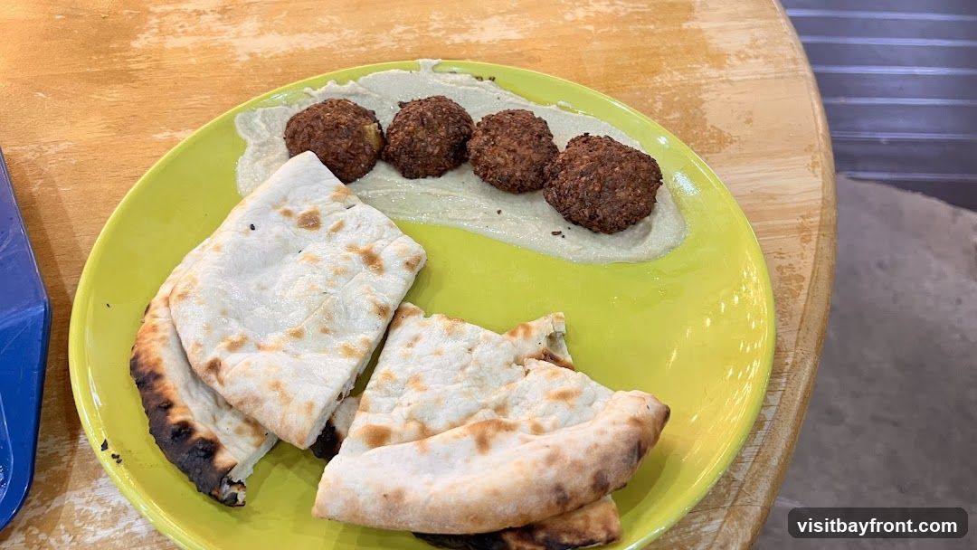 A bright yellow plate holds a serving of hummus topped with four falafel balls. Several pieces of flatbread, some with slightly charred edges, are arranged on the side of the plate. The plate sits on a wooden table.