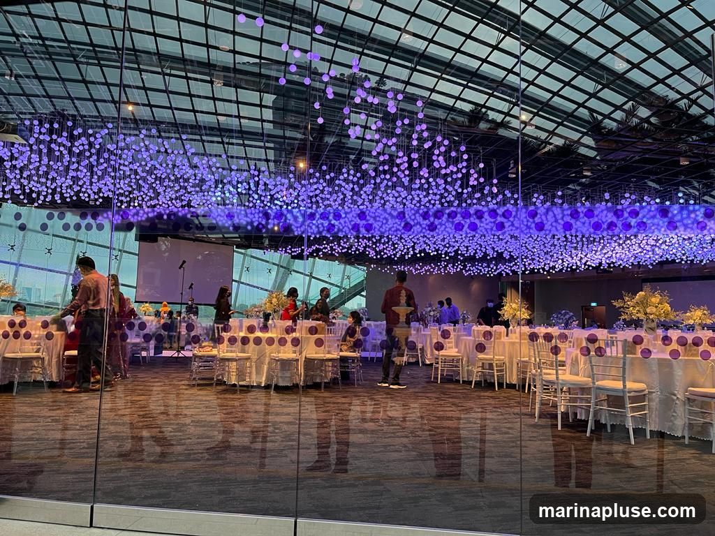 A large event hall is set up with round tables draped in white tablecloths, each adorned with purple circular decorations. White chairs are arranged around the tables. The ceiling is a glass structure with a grid pattern, from which numerous strings of small purple lights and larger purple orbs are suspended, creating a dazzling effect. In the background, a projector screen is visible, along with several people attending to the setup or guests. The floor is reflective, mirroring the lights and tables.