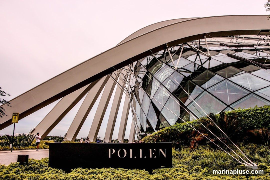 A large, geodesic dome structure with a white, curved roof supported by angled beams. The dome is made of glass panels in a diamond pattern. In the foreground, a black sign with the word "POLLEN" in white capital letters is visible, surrounded by lush green foliage. People can be seen walking in the background near the entrance of the dome.